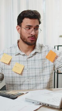 Tired exhausted fatigue young Indian businessman office employee manager sits in home office table with laptop sticky notes with many tasks inscription help, feels sad boredom overworked multitasking.