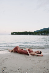 Relaxed woman in rust dress lying on sandy beach by calm sea water with distant palm trees and hills under soft sky at sunset.