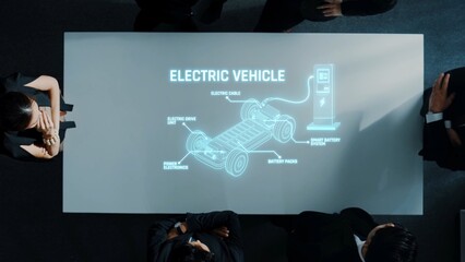 A professional team engages in a strategic discussion about electric vehicle technology. The meeting features a detailed projection of vehicle components and charging solutions. Raster