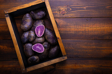 Obraz premium Harvest of ripe purple potatoes, unusual raw potatoes in the box on wooden background top view