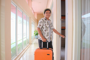 Asian traveler in hotel corridor pulling orange suitcase, wearing tropical patterned shirt and hat. Bright modern hotel background enhances the travel and tourism theme. © Rudy
