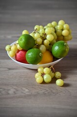 A white bowl overflowing with fresh fruit, including crisp green grapes, bright green limes, and a red apple, sits on a wooden table.