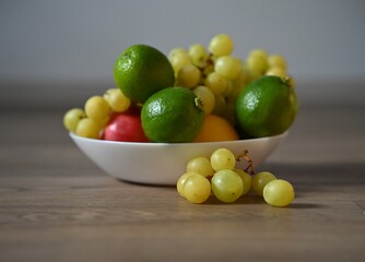 A shot from a lower angle of a white bowl filled with green grapes, limes, and an apple on a wooden floor. Several grapes are scattered outside the bowl, emphasising the freshness and natural feel.