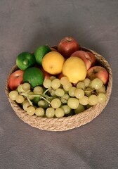 A woven basket filled with fresh, healthy fruits, including green grapes, red apples, vibrant lemons, and green limes, sits on a grey surface, perfectly suited for a balanced diet.
