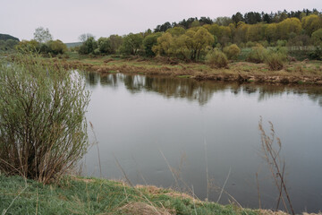 Tranquil spring river landscape with calm water, green shore grasses and reflections of trees, serene nature scenery in rural Russia, peaceful countryside atmosphere