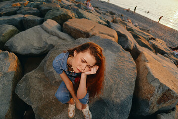 Fototapeta premium lifestyle woman in modern boho-western denim street style sitting on rocks at beach with warm film color tones, thoughtful mood, casual summer outfit