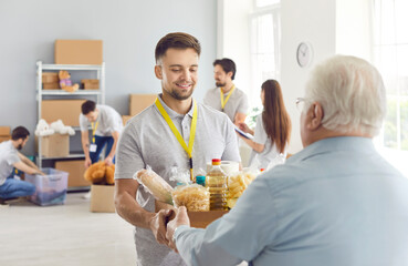 Young smiling friendly male volunteer gives a box full of grocery goods to an aged, mature, retired...