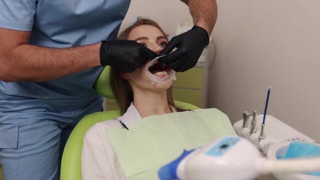 Dental examination, Healthy teeth, Dental therapy. Dentist in blue scrubs examines woman's teeth, who is sitting in dental chair with mouth retractor in place.