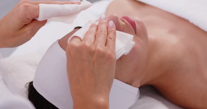 Close-up photo pf professional cosmetologist or beautician doctor hands making anti-age procedures cleaning with napkins facial mask of young pretty woman client in beauty spa clinic. 4k video.
