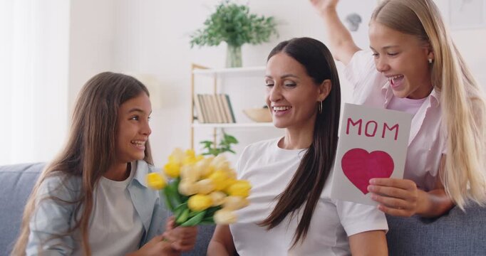 Happy mothers day surprise smiling loving teen daughter sisters greeting happy mother with anniversary presenting bouquet of flowers, card, happy mom and grownup children in hug embraces to celebrate 