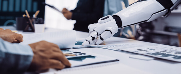 A futuristic office scene showcases a robotic arm engaging with human hands over documents and graphs, highlighting productivity and collaboration in a digital workspace.Noogenesis