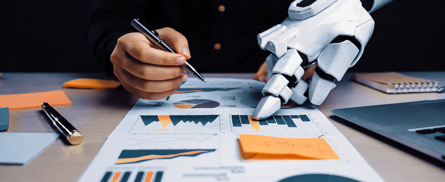 A unique scene depicting a robotic hand assisting a human in analyzing business charts and graphs, showcasing the future of technology and productivity in the workplace.Noogenesis