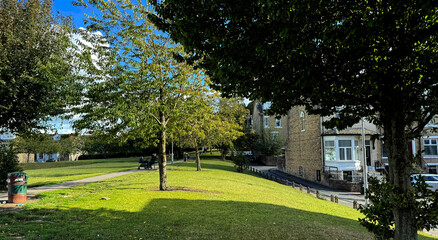 A peaceful park scene captures the afternoon sunlight casting shadows across the grass. Trees with full foliage outline a pathway while quaint brick houses flank the park on the right in, Bradford, UK
