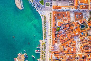 Drone top down image of Split Riva showing waterfront promenade, palm trees, stone buildings, cafes, docked boats and clear Adriatic waters in the historic Dalmatian city