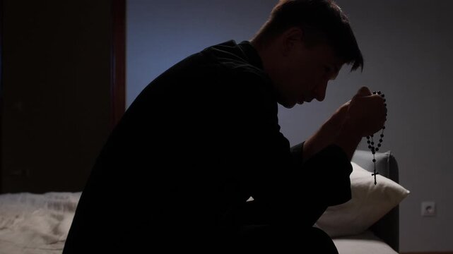 Young priest praying with rosary in dark room