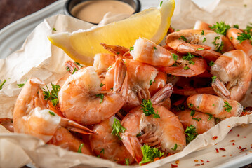 Cooked shrimp served on parchment with creamy dipping sauce, lemon wedge, parsley garnish, and red pepper flakes. Clean, appetizing seafood presentation on white plate.