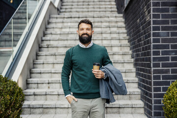 Portrait of smiling smart casual executive standing near staircase with coffee to go in hands and looking at camera.