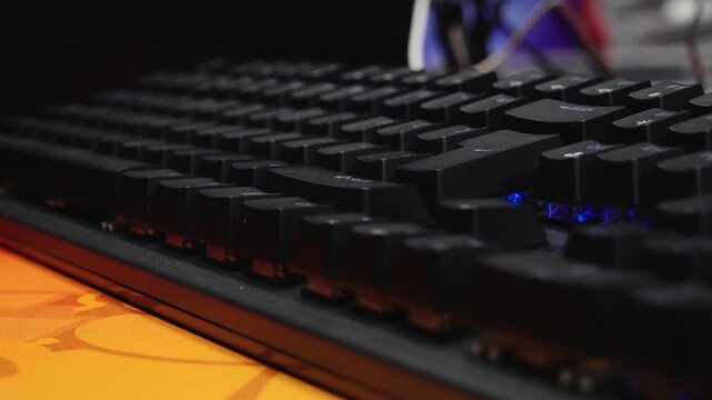 Close-up view of a black mechanical gaming keyboard with raised keycaps and backlit keys on an orange mousepad in a tech or gaming environment.
