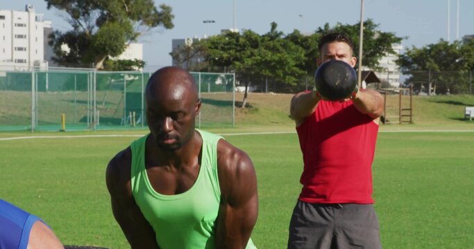Men wearing neon tank top and red shirt swinging black kettlebells on grassy sports field - Powered by Adobe