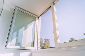An open white plastic window in a bright, minimalist room, letting in sunlight and a summer breeze. The serene view from the apartment looks out on a clear blue sky and green trees.