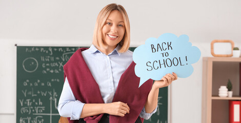 Mature female math teacher holding paper sheet with text BACK TO SCHOOL in classroom
