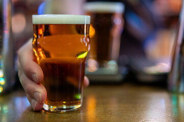 Close-up of a pint of amber beer with frothy head held by hand on wooden bar table, with blurred background glass and warm pub lighting. Casual, social atmosphere.