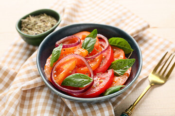 Bowl of fresh tomato salad with red onion and basil leaves on beige wooden background, closeup