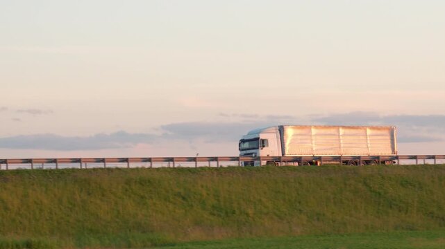 Powerful modern semi trailers truck is moving along the expressway at sunset. Lorry driving through green hills and metal barriers on intercity highway. Cargo transportation goods