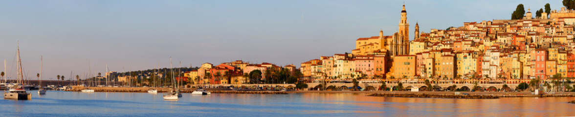 Obraz premium Panoramic sunrise view of Menton, France, with colorful old town buildings glowing in warm light, set against the Mediterranean Sea and blue sky on the French Riviera