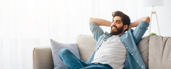 Home comfort. Young arab man relaxing on cozy sofa in living room, leaning back on couch with hands...