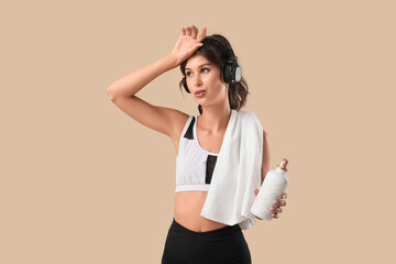 Tired sporty young woman with headphones, water bottle and towel on beige background