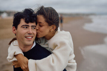 Romantic young couple enjoying a playful piggyback ride on the beach, with the woman sweetly kissing the man on the cheek
