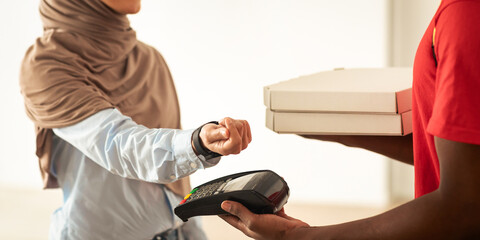 Smart Payment, Food Delivery. Smiling Muslim Woman In Hijab Paying For Pizza With Smartwatch Receiving Box From Courier Guy Standing At Door. Restaurant Delivering Service. Selective Focus On Hand