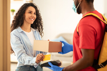 Payment Upon Receipt. Smiling Client Paying With Debit Card For Parcel Receiving Box From Black Courier Guy In Protective Gloves And Mask Standing At Home Door. Quick Express Delivering Service