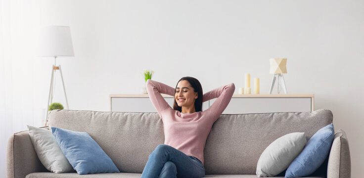 Stay at home alone, enjoy silence, positive, rest and relax in your own cozy apartment after cleaning. Serene happy arab pretty woman sits on sofa in minimalist living room and looking at empty space