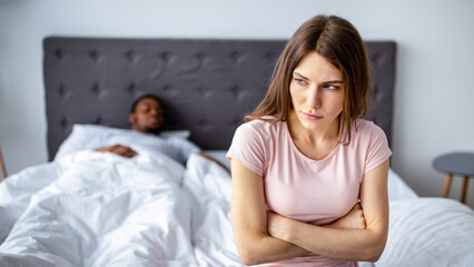 Depressed young woman having relationship problem, sitting on bed while her husband sleeping at...