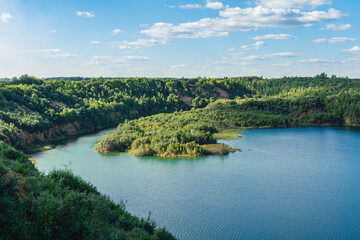 Fototapeta premium A serene river winding through lush green forested hills under a bright blue sky with scattered clouds, showcasing peaceful nature and vibrant landscapes.