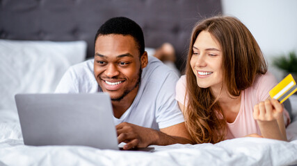 Online shopping concept. Cheerful interracial couple with credit card and laptop pc buying goods in internet store on bed at home. Joyful black guy and his wife purchasing things remotely