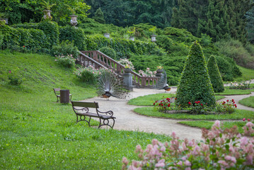 A picturesque garden with neatly trimmed bushes, blooming flowers, benches, and two peacocks...