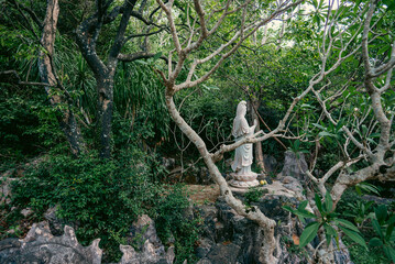 Serene marble statue of Guanyin in forest shrine with offerings at the base, Peaceful Buddhist female figure in jungle setting with altar and sacred atmosphere