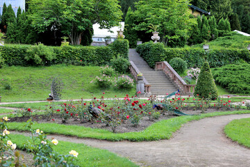 A picturesque garden with neatly trimmed bushes, blooming flowers, benches, and peacocks strolling among the flowers near the stone staircase.
