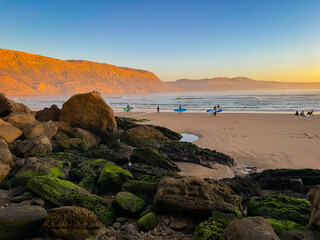 Sandy beach at sunset, with large rocks covered with green algae in foreground. Several surfers carrying their surfboards are walking along sandy shore, while hill in background glows in golden shade. © helivideo