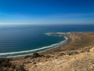 Sweeping curve of coastline near the village of Imsouane. Blue Atlantic Ocean meets sandy beach, with a few waves breaking perfectly near the famous and popular surf town at the end of a small cape.