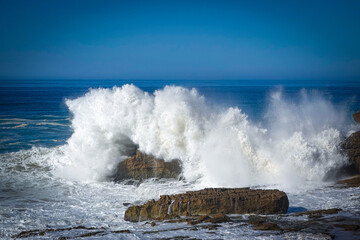 Massive and powerful wave crashes with immense force against a rugged cliff and surrounding rocks, creating a dramatic explosion of white water and foam. Dramatic moment when ocean hits the land.