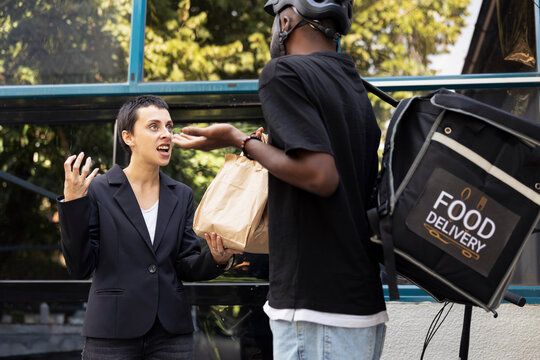 Unhappy customer refuses a takeout meal from black biker employee with a courier bag. Dissatisfied woman gestures in protest for the wrong fast food order, demanding a refund.