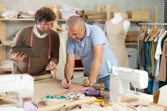 Adult male tailor showing elderly male assistant pattern drawing on tablet in sewing workshop