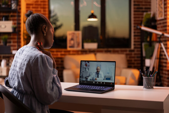 Female entrepreneur sits at home during online medical consultation on laptop, speaking with doctor. Woman attends virtual healthcare appointment, receiving professional health advice from physician.