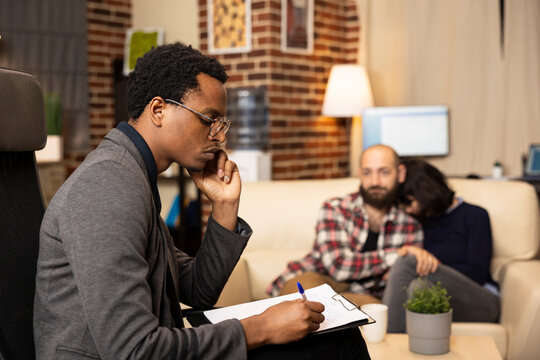 Thoughtful psychiatrist holds chin and takes notes as loving couple waits for relationship assessment results. Black therapist writes on clipboard, focused on evaluating couple during therapy session.