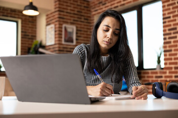 Indian female professional taking notes by hand beside laptop in brick wall apartment. Concentrated businesswoman outlining strategic business plans in notepad at home office desk.