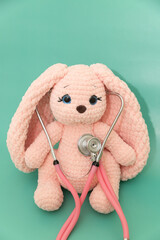 Pink plush knitted bunny listening to heart with pink stethoscope on green background, playing doctor